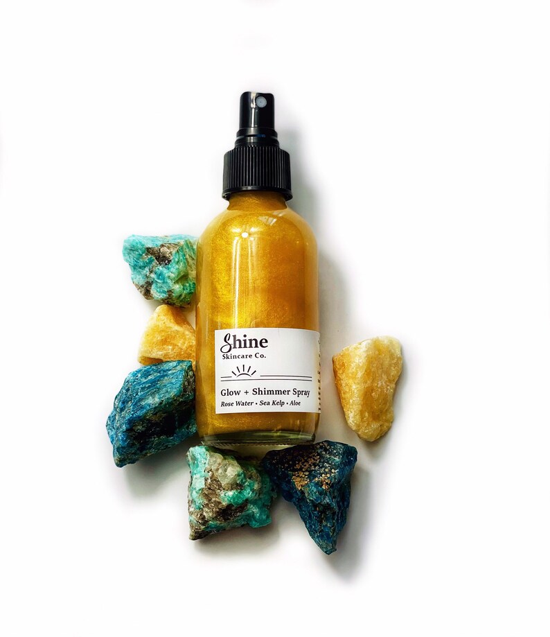 May include: A clear glass bottle filled with a golden liquid, labeled "Shine Skincare Co. Glow + Shimmer Spray." The bottle is surrounded by colorful, rough-cut gemstones in shades of blue, turquoise, and yellow. The spray bottle has a black nozzle.