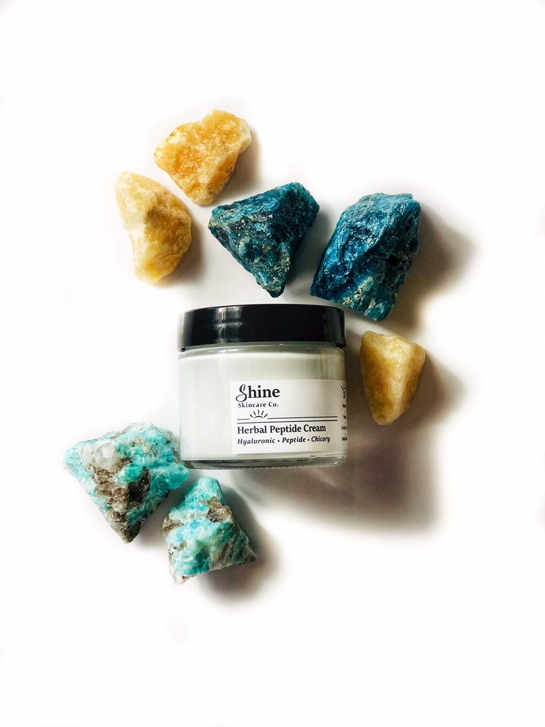 Organic skincare anti-aging face moisturizer in clear glass jar with black lid. Face cream is a very pale blue color from blue tansy oil. Photo of jar on white background surrounded by blue and yellow crystal gemstones.