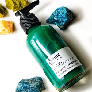 Puede incluir: Una botella de vidrio verde con una tapa de bomba negra y una etiqueta blanca que dice "Shine Skincare Co. Greens + Ginseng Cleanser Chlorophyll, Green Tea, Spirulina".