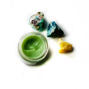 May include: A small glass jar with a green balm inside, surrounded by four colorful gemstones. The gemstones are green, blue, and yellow.
