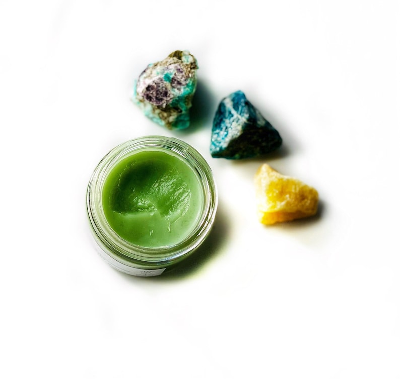 May include: A small glass jar with a green balm inside, surrounded by four colorful gemstones. The gemstones are green, blue, and yellow.