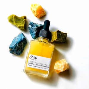May include: A clear glass bottle of serum with a black dropper. The bottle is labeled "Shine Skincare Co. Soothe + Hydrate Serum Protein - Hyaluronic Acid - Algae". The bottle is surrounded by yellow, green, and blue crystals.