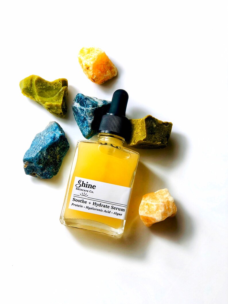 May include: A clear glass bottle of serum with a black dropper. The bottle is labeled "Shine Skincare Co. Soothe + Hydrate Serum Protein - Hyaluronic Acid - Algae". The bottle is surrounded by yellow, green, and blue crystals.