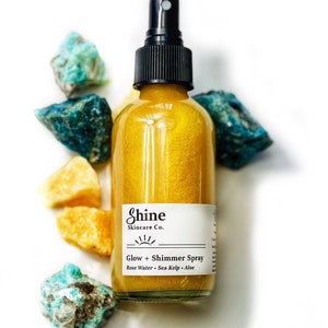 May include: A clear glass bottle of Shine Skincare Co. Glow + Shimmer Spray with a black spray nozzle. The bottle contains a golden liquid and is surrounded by blue and yellow gemstones. The label lists Rose Water, Sea Kelp, and Aloe.
