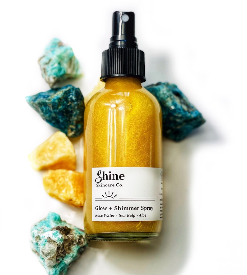May include: A clear glass bottle of Shine Skincare Co. Glow + Shimmer Spray with a black spray nozzle. The bottle contains a golden liquid and is surrounded by blue and yellow gemstones. The label lists Rose Water, Sea Kelp, and Aloe.