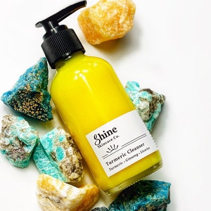 Organic skincare turmeric cleanser in clear glass bottle with pump dispenser. Face wash is bright yellow color and photographed on white background.  Skincare product is along side blue and yellow crystals.