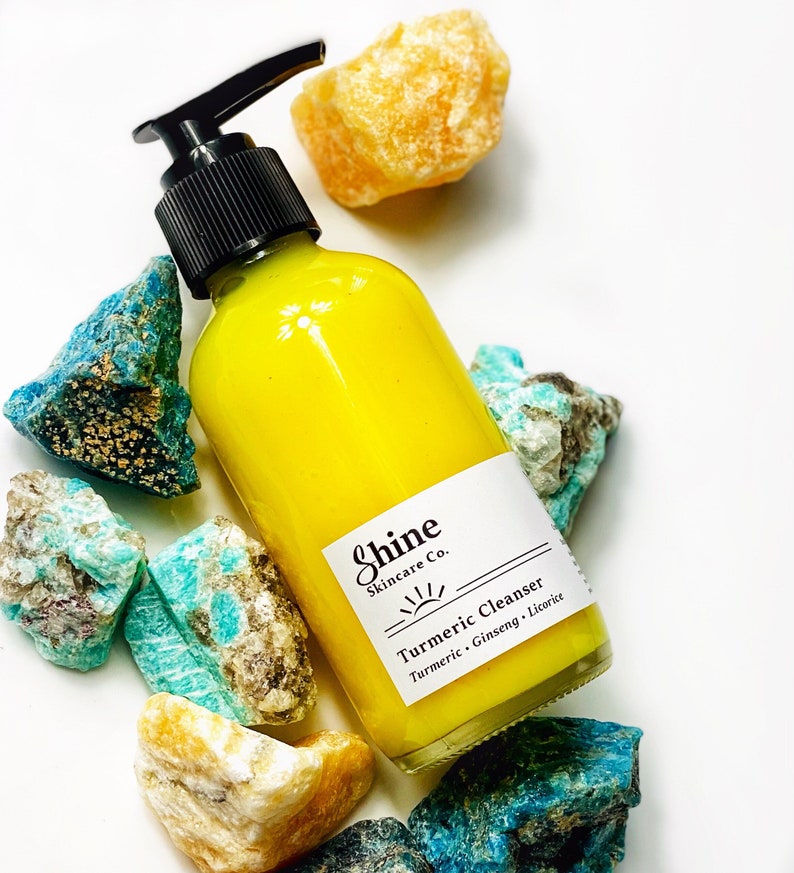 Organic skincare turmeric cleanser in clear glass bottle with pump dispenser. Face wash is bright yellow color and photographed on white background.  Skincare product is along side blue and yellow crystals.