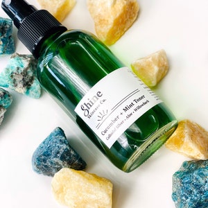 May include: A green glass bottle with a black spray top filled with a clear liquid. The bottle has a white label with the text "Shine Skincare Co." and "Cucumber + Mint Toner Colloidal Silver - Aloe - Willowbark". The bottle is surrounded by yellow and blue crystals.