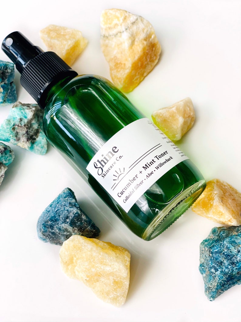 May include: A green glass bottle with a black spray top filled with a clear liquid. The bottle has a white label with the text "Shine Skincare Co." and "Cucumber + Mint Toner Colloidal Silver - Aloe - Willowbark". The bottle is surrounded by yellow and blue crystals.