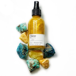 May include: A gold-colored shimmer spray bottle with a black spray nozzle, surrounded by colorful gemstones. The bottle has a white label with the text "Shine Skincare Co." and "Glow + Shimmer Spray". The ingredients listed are Rose Water, Sea Kelp, and Aloe.