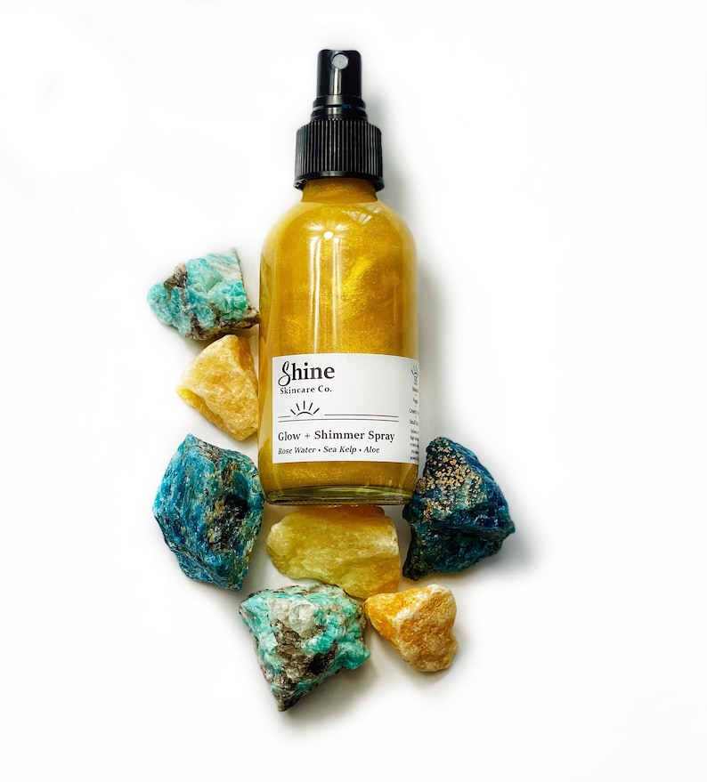 May include: A gold-colored shimmer spray bottle with a black spray nozzle, surrounded by colorful gemstones. The bottle has a white label with the text "Shine Skincare Co." and "Glow + Shimmer Spray". The ingredients listed are Rose Water, Sea Kelp, and Aloe.