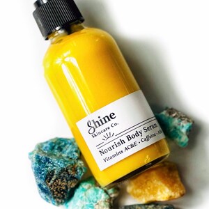 Puede incluir: Una botella de s&eacute;rum corporal amarillo con una bomba negra y una etiqueta blanca que dice "Shine Skincare Co. Nourish Body Serum Vitamins AC&E Caffeine + AHA". La botella est&aacute; rodeada de piedras azules y amarillas.