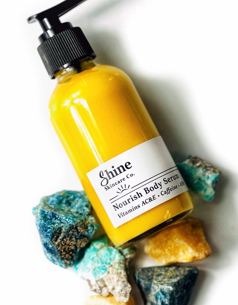 Puede incluir: Una botella de s&eacute;rum corporal amarillo con una bomba negra y una etiqueta blanca que dice "Shine Skincare Co. Nourish Body Serum Vitamins AC&E Caffeine + AHA". La botella est&aacute; rodeada de piedras azules y amarillas.
