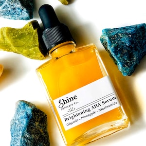 Organic skincare product brightening face serum in clear square glass bottle. Serum surrounded by yellow, blue and green crystal gemstones on white background. Shine Skincare label reads brightening aha serum with glycolic, pineapple and niacinamide