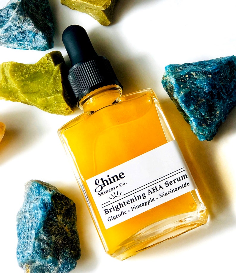 Organic skincare product brightening face serum in clear square glass bottle. Serum surrounded by yellow, blue and green crystal gemstones on white background. Shine Skincare label reads brightening aha serum with glycolic, pineapple and niacinamide