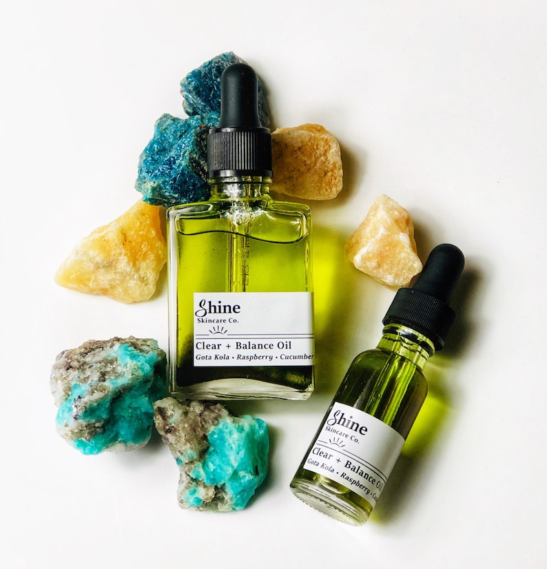 May include: Two glass bottles of green liquid with dropper tops. Both bottles have a white label with the text "Shine Skincare Co." and "Clear + Balance Oil" with a list of ingredients. The bottles are sitting on a white surface with green and yellow crystals.