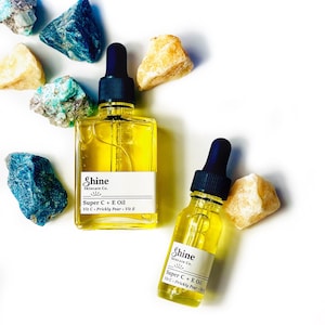 May include: Two glass bottles of Shine Skincare Co. Super C + E Oil with black droppers, filled with yellow liquid. The bottles are surrounded by colorful rocks on a white background. The labels read "Super C + E Oil Vit C + Prickly Pear + Vit E."