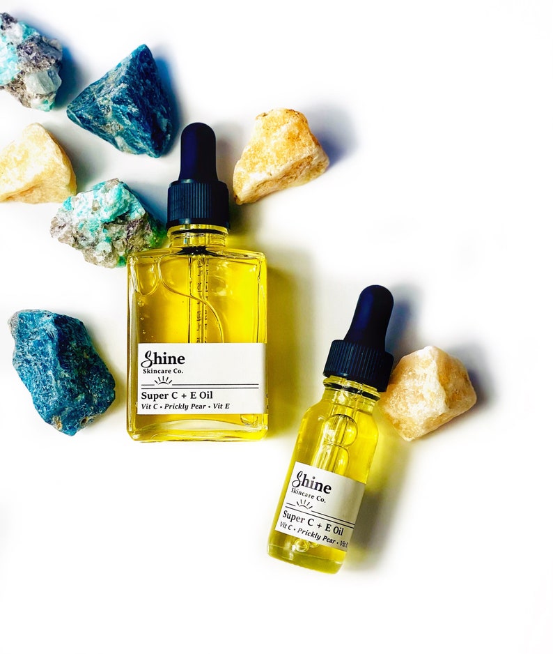 May include: Two glass bottles of Shine Skincare Co. Super C + E Oil with black droppers, filled with yellow liquid. The bottles are surrounded by colorful rocks on a white background. The labels read "Super C + E Oil Vit C + Prickly Pear + Vit E."