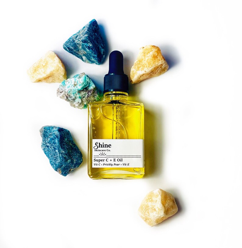 May include: A square glass bottle of Shine Skincare Co. Super C + E Oil with a dark blue dropper top. The bottle is surrounded by blue, green, and yellow-beige rough-cut stones on a white background. The label includes the text "Vit C + Prickly Pear + Vit E."