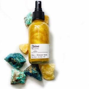 May include: A gold shimmer spray bottle with a black spray nozzle, labeled "Shine Skincare Co." and "Glow + Shimmer Spray." The bottle is surrounded by blue and yellow-toned rough-cut stones, all set against a white background.