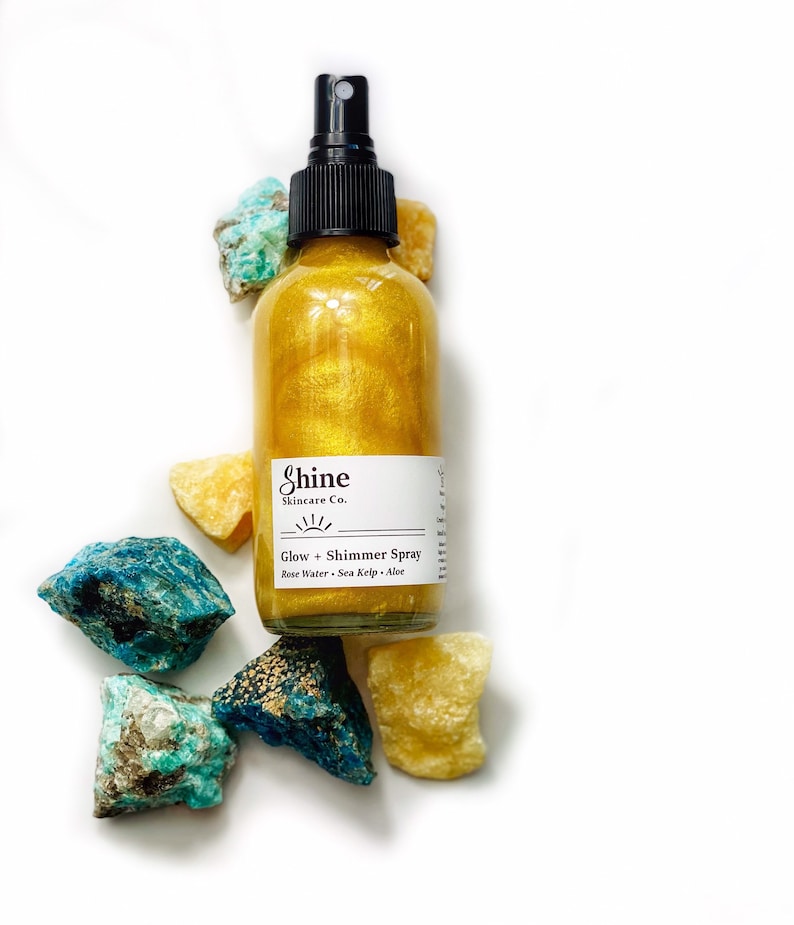 May include: A gold shimmer spray bottle with a black spray nozzle, labeled "Shine Skincare Co." and "Glow + Shimmer Spray." The bottle is surrounded by blue and yellow-toned rough-cut stones, all set against a white background.