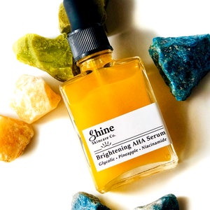 May include: A glass bottle of yellow serum with a black dropper. The bottle is labelled "Shine Skincare Co. Brightening AHA Serum Glycolic - Pineapple - Niacinamide". The bottle is surrounded by green, yellow, and blue stones.