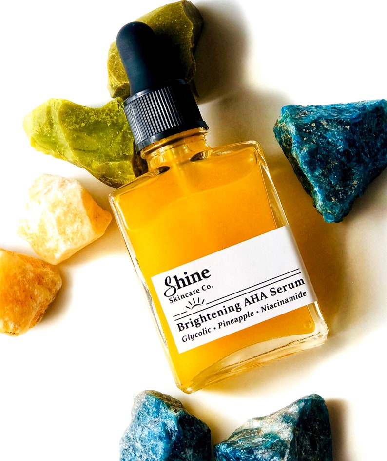 May include: A glass bottle of yellow serum with a black dropper. The bottle is labelled "Shine Skincare Co. Brightening AHA Serum Glycolic - Pineapple - Niacinamide". The bottle is surrounded by green, yellow, and blue stones.