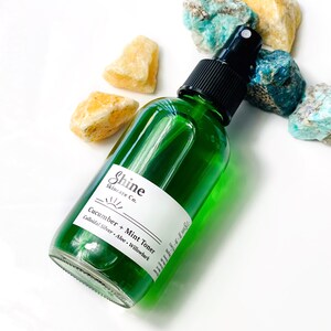 May include: A green glass bottle with a black spray top, labeled "Shine Skincare Co. Cucumber + Mint Toner Colloidal Silver, Aloe, Willowbark".