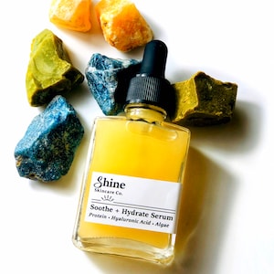 May include: A clear glass bottle with a yellow liquid inside. The bottle has a black dropper top and a white label that says "Shine Skincare Co." and "Soothe + Hydrate Serum". The label also lists the ingredients: "Protein - Hyaluronic Acid - Algae". The bottle is surrounded by colorful gemstones.