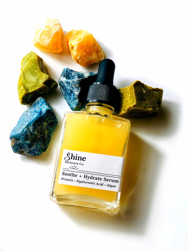 May include: A clear glass bottle with a yellow liquid inside. The bottle has a black dropper top and a white label that says "Shine Skincare Co." and "Soothe + Hydrate Serum". The label also lists the ingredients: "Protein - Hyaluronic Acid - Algae". The bottle is surrounded by colorful gemstones.