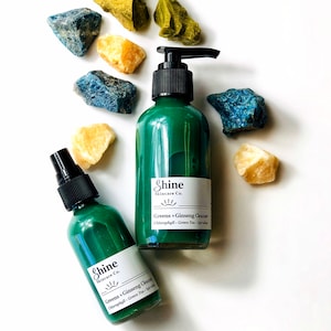 Natural skincare green tea cleanser in clear glass bottle with pump lid. Face wash is a dark green color and thick consistency. Two bottles in photo with white background with blue, green and yellow crystal gemstones around top of bottles.