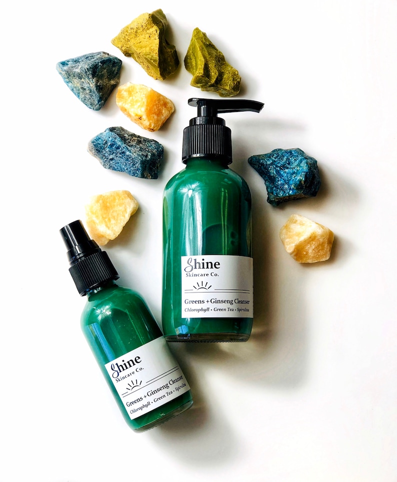 Natural skincare green tea cleanser in clear glass bottle with pump lid. Face wash is a dark green color and thick consistency. Two bottles in photo with white background with blue, green and yellow crystal gemstones around top of bottles.