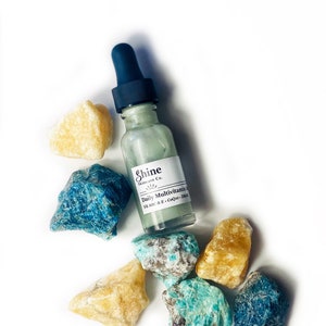 Op de afbeelding: Een heldere glazen fles met een druppelaar, gevuld met een lichtgroene vloeistof. De fles is gelabeld met "Shine Skincare Co. Daily Multivitamin Serum Vit A,B,C & E, CoQ10, DMAE". De fles is omringd door verschillende groene en gele stenen.