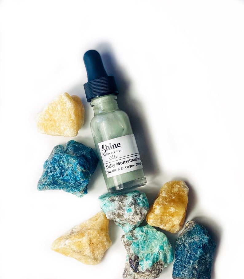 Op de afbeelding: Een heldere glazen fles met een druppelaar, gevuld met een lichtgroene vloeistof. De fles is gelabeld met "Shine Skincare Co. Daily Multivitamin Serum Vit A,B,C & E, CoQ10, DMAE". De fles is omringd door verschillende groene en gele stenen.