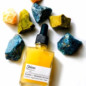 May include: A clear glass bottle with a yellow liquid inside. The bottle has a white label with the text "Shine Skincare Co." and "Soothe + Hydrate Serum" in black. The label also lists the ingredients: "Protein - Hyaluronic Acid - Algae". The bottle is surrounded by colorful stones.