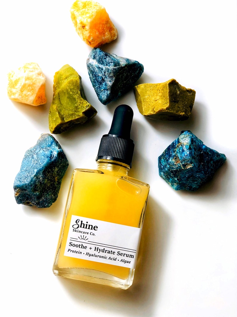 May include: A clear glass bottle with a yellow liquid inside. The bottle has a white label with the text "Shine Skincare Co." and "Soothe + Hydrate Serum" in black. The label also lists the ingredients: "Protein - Hyaluronic Acid - Algae". The bottle is surrounded by colorful stones.