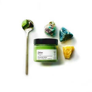 May include: A green cleansing balm jar with a black lid and white label that says "Shine Skincare Co. Cleansing Balm Matcha - Prickly Pear - Mulberry" with a sun and a star symbol. The jar is on a white surface with green and blue stones and a silver spoon.