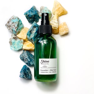 Organic skincare cucumber toner in clear glass bottle with black spray top. Face toner is bright green in color. Photo taken on white background with blue and yellow crystal gemstones along the side of the product.