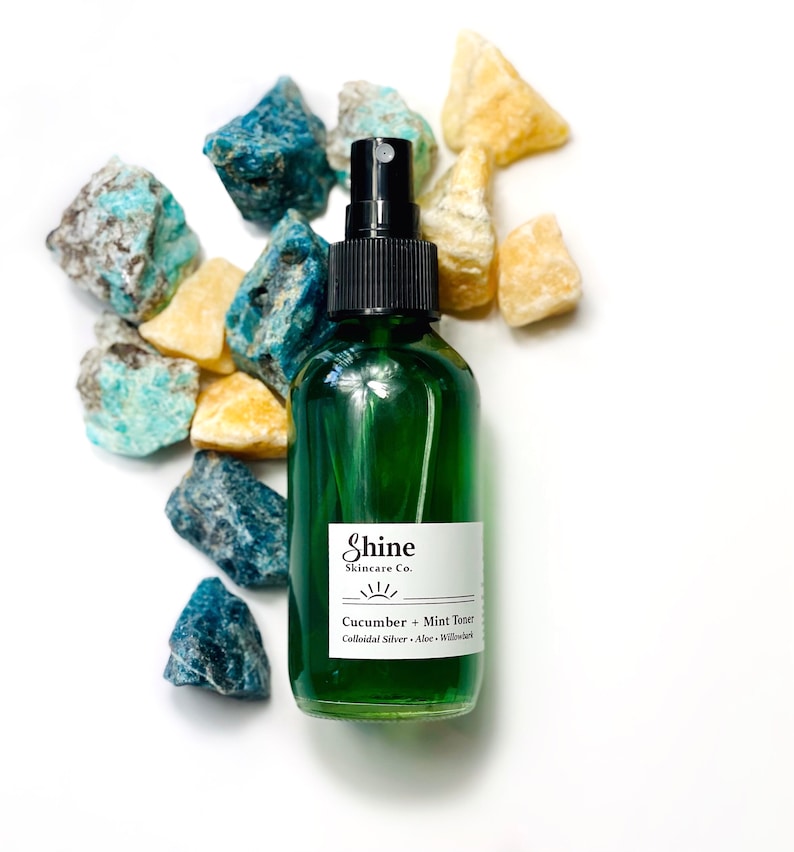 Organic skincare cucumber toner in clear glass bottle with black spray top. Face toner is bright green in color. Photo taken on white background with blue and yellow crystal gemstones along the side of the product.