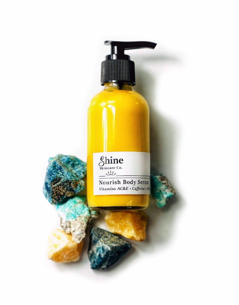 Puede incluir: Una botella de s&eacute;rum corporal amarillo con una bomba negra, etiquetada como "Nourish Body Serum" por Shine Skincare Co. La botella est&aacute; rodeada de piedras de colores en bruto en tonos de azul, turquesa y oro, sobre un fondo blanco.