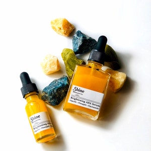 May include: Two amber glass bottles with dropper tops filled with a yellow liquid. The bottles are labelled "Shine Skincare Co. Brightening AHA Serum" and "Shine Skincare Co. Brightening AHA". The bottles are surrounded by yellow, green, and blue crystals.