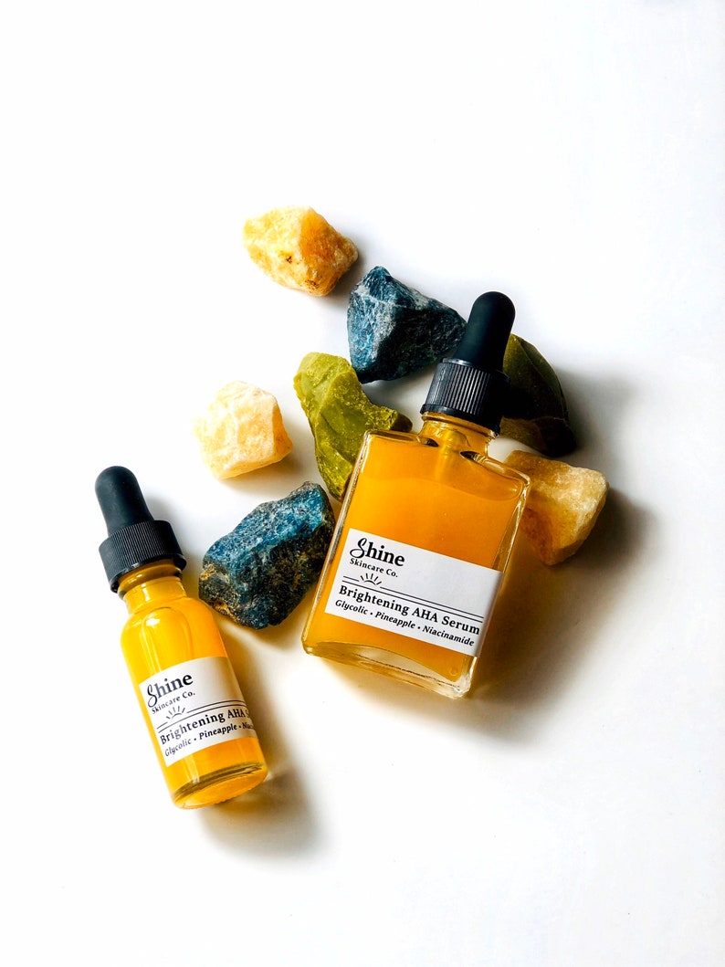 May include: Two amber glass bottles with dropper tops filled with a yellow liquid. The bottles are labelled "Shine Skincare Co. Brightening AHA Serum" and "Shine Skincare Co. Brightening AHA". The bottles are surrounded by yellow, green, and blue crystals.