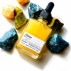 May include: A clear glass bottle with a yellow liquid inside. The bottle has a black dropper top and a white label that reads "Shine Skincare Co. Soothe + Hydrate Serum Protein, Hyaluronic Acid, Algae". The bottle is surrounded by small blue, green, and yellow rocks.