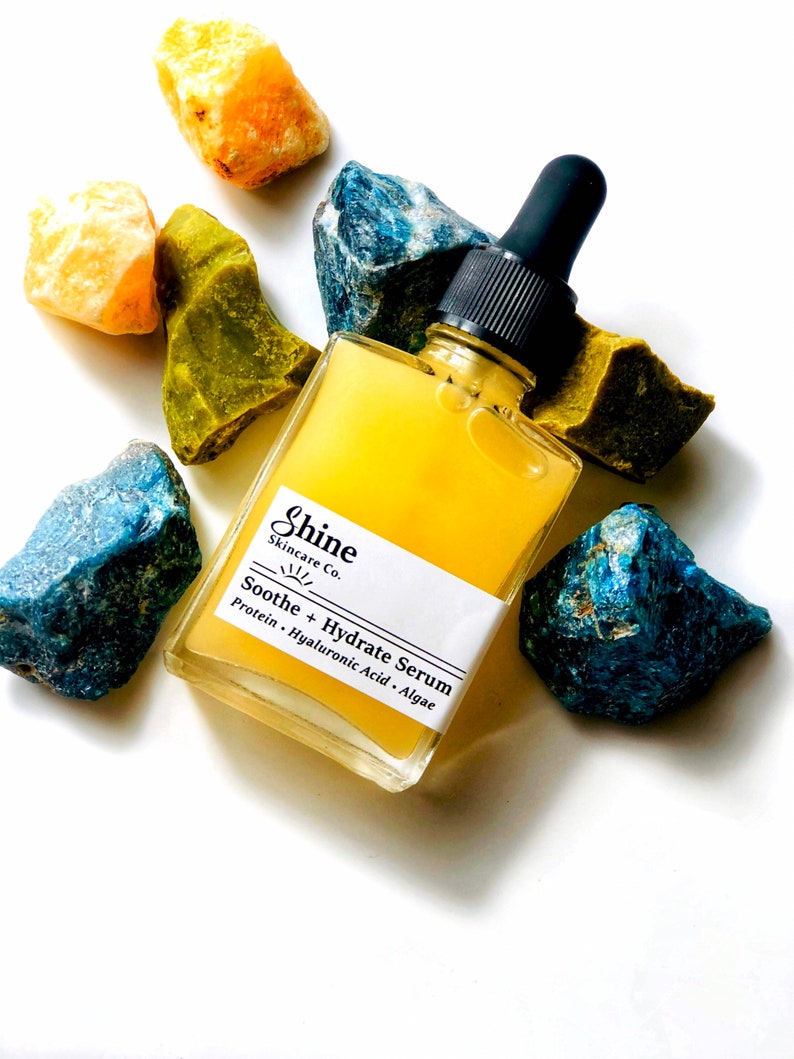 May include: A clear glass bottle with a yellow liquid inside. The bottle has a black dropper top and a white label that reads "Shine Skincare Co. Soothe + Hydrate Serum Protein, Hyaluronic Acid, Algae". The bottle is surrounded by small blue, green, and yellow rocks.