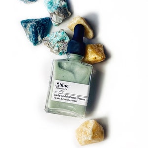 Op de afbeelding: Een doorzichtige glazen fles met een groene serum. De fles heeft een zwarte druppelaar en een wit etiket met de tekst "Shine Skincare Co. Daily Multivitamin Serum Vit A B C & E - CoQ10 - DMAE". De fles is omringd door kleine, lichtblauwe en gele stenen.