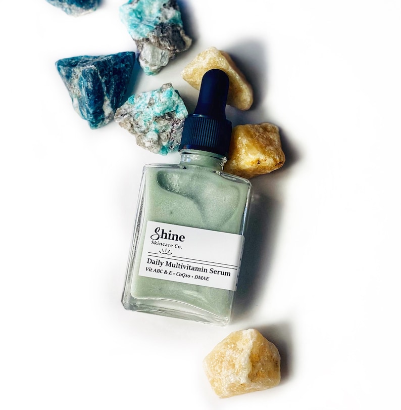 Op de afbeelding: Een doorzichtige glazen fles met een groene serum. De fles heeft een zwarte druppelaar en een wit etiket met de tekst "Shine Skincare Co. Daily Multivitamin Serum Vit A B C & E - CoQ10 - DMAE". De fles is omringd door kleine, lichtblauwe en gele stenen.