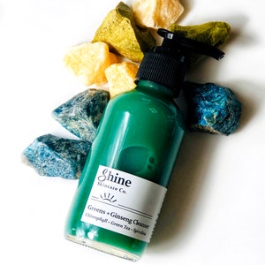 Puede incluir: Una botella de vidrio verde con una tapa de bomba negra y una etiqueta blanca que dice "Shine Skincare Co. Greens + Ginseng Cleanser Chlorophyll, Green Tea, Spirulina"