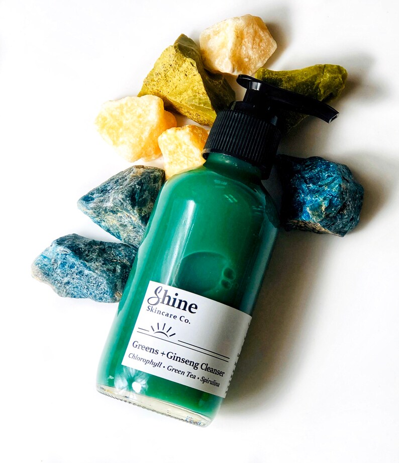 Puede incluir: Una botella de vidrio verde con una tapa de bomba negra y una etiqueta blanca que dice "Shine Skincare Co. Greens + Ginseng Cleanser Chlorophyll, Green Tea, Spirulina"