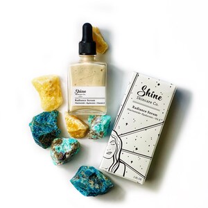 May include: A glass bottle of Shine Skincare Co. Radiance Serum with a dropper, sitting on a white surface with blue and yellow crystals. The bottle label reads "Shine Skincare Co. Radiance Serum Niacinamide Hyaluronic Vit E".