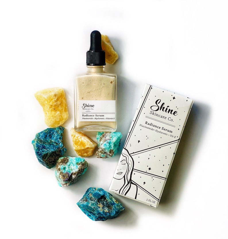 May include: A glass bottle of Shine Skincare Co. Radiance Serum with a dropper, sitting on a white surface with blue and yellow crystals. The bottle label reads "Shine Skincare Co. Radiance Serum Niacinamide Hyaluronic Vit E".